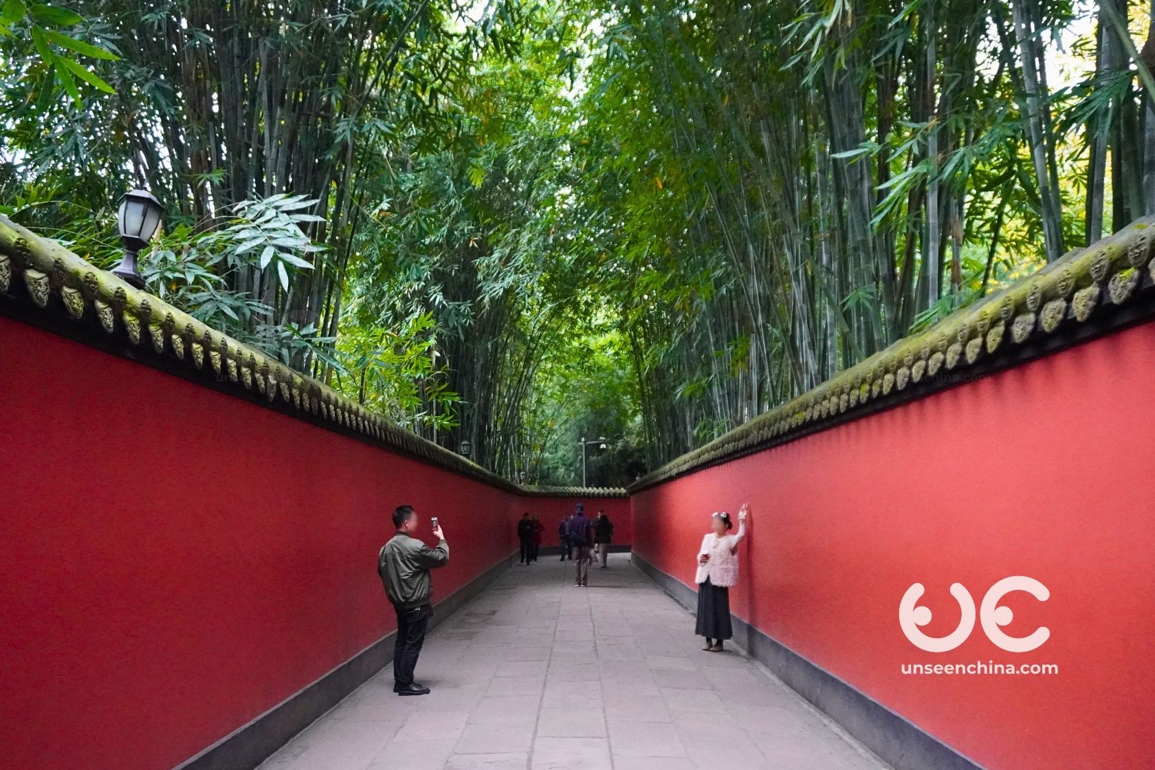 A straight section of the Wuhou Shrine's iconic red wall serves as a backdrop for a couple posing for a picture, shaded by bamboo trees.